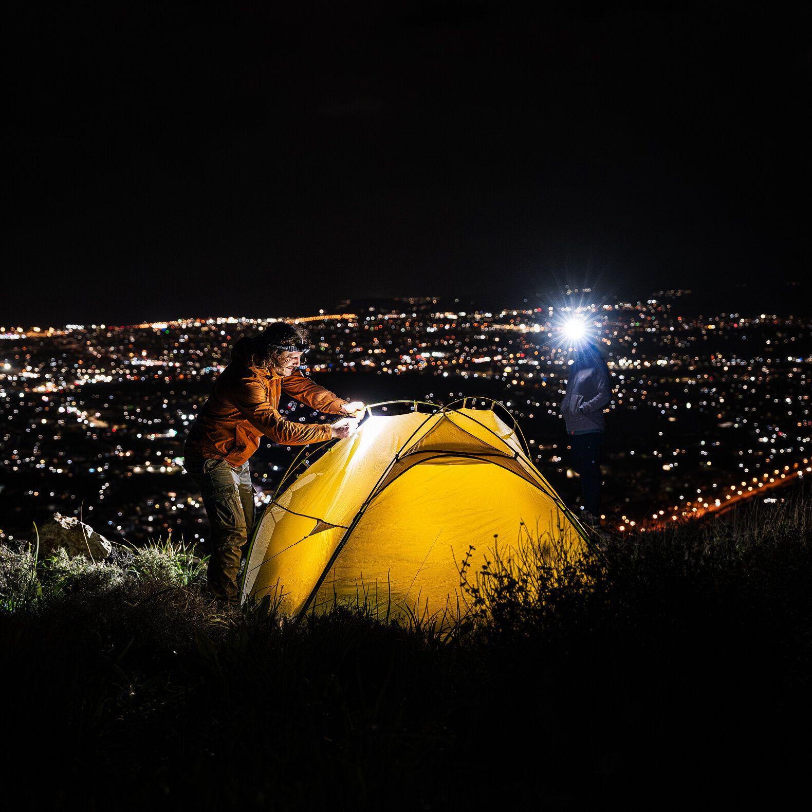 Zwei Personen bauen nachts ein gelbes Campingzelt auf einem Hügel mit Blick auf eine Stadt auf, mit hellen Scheinwerfern zur Beleuchtung.