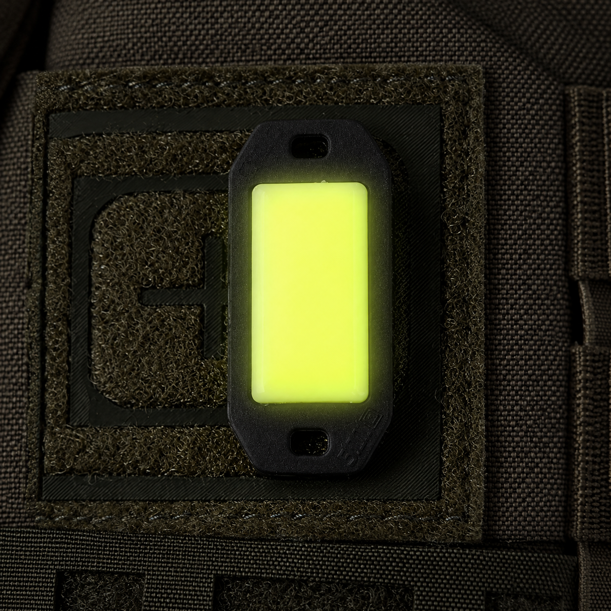 Ein im Dunkeln leuchtender 5.11 Tactical-Markierungsfleck, der am olivfarbenen MOLLE-Gurtband befestigt ist, um die Notfallausrüstung zu identifizieren.
