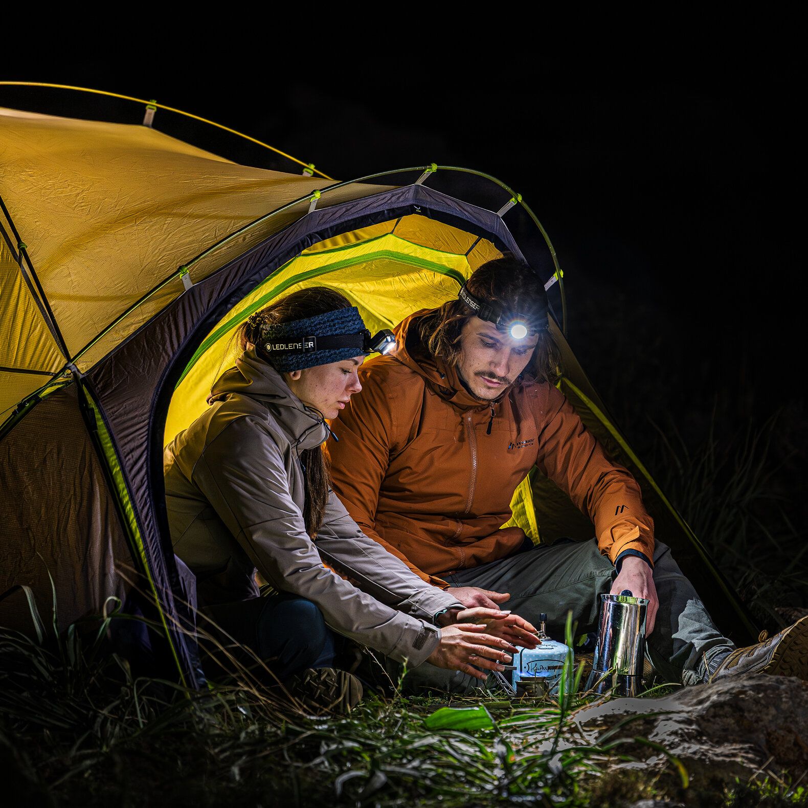 Zwei Personen mit Ledlenser-Stirnlampen campen nachts und nutzen einen tragbaren Ofen in einem gelben Zelt, um sich zu wärmen.