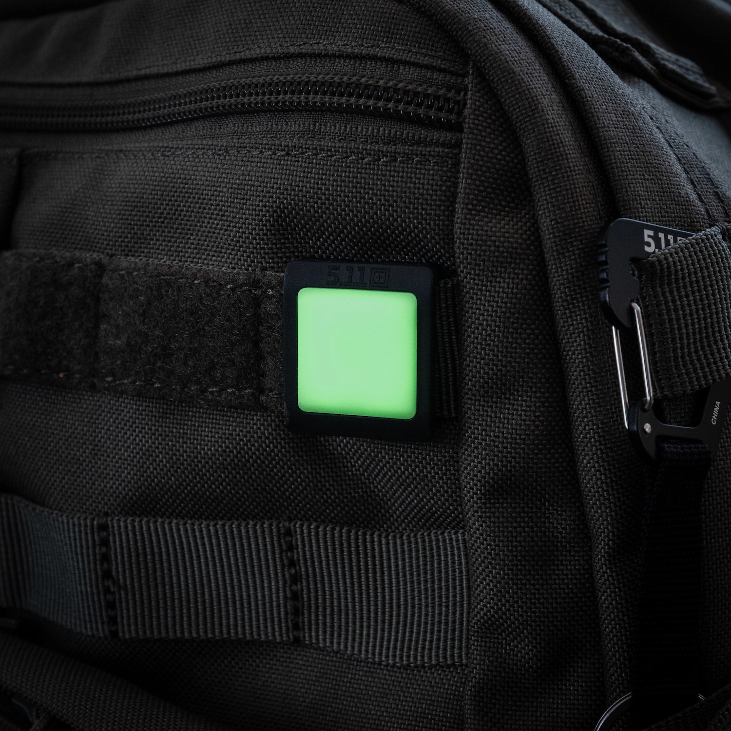 Ein quadratischer 5.11 Tactical-Marker, der im Dunkeln leuchtet und an einem schwarzen MOLLE-Taktikrucksack befestigt ist, um im Notfall sichtbar zu sein.